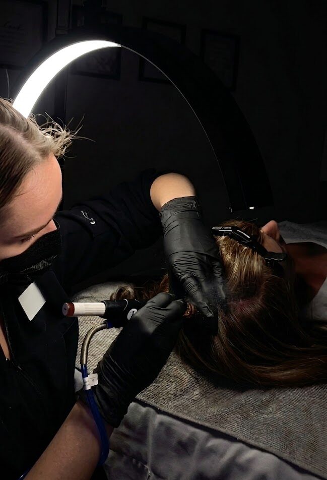 Practitioner uses a device on a person’s scalp under a ring light while the person lies on a treatment bed