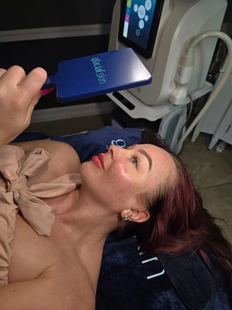 Person reclining on a treatment bed while holding a mirror near a skincare device.