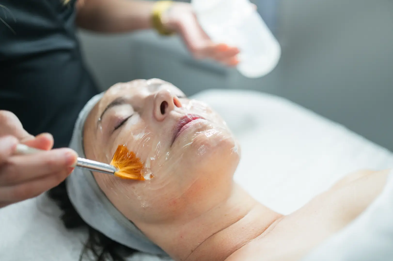 Esthetician applying a facial mask to a reclining patient with a brush